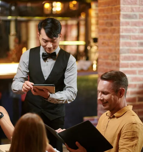 Waiter in a bow tie taking an order on a tablet from guests at a restaurant table