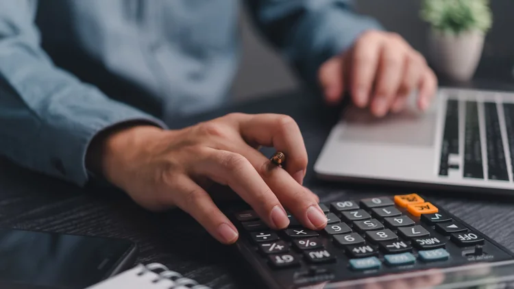 Person using a calculator next to a laptop while reviewing payroll figures